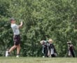 Young golfer swinging a club on the course, with golf bags and clubs in the background amid lush greenery.