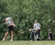 A golfer swings a club on the course, with another player and golf bags in the background amidst a sunny landscape.