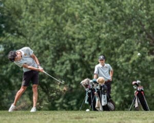 A golfer swings a club on the course, with another player and golf bags in the background amidst a sunny landscape.