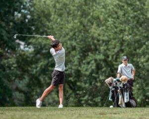 A golfer swings on the course, while a caddie carries golf bags in the background, surrounded by lush greenery.