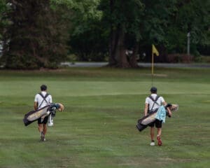 Two golfers walk down a lush fairway carrying golf bags, heading towards the green with a flag in the background.
