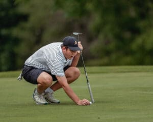 Young golfer kneels on the green, preparing to putt, focused on the ball with a putter in hand.
