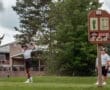 Young golfer swinging a club on the course, with fellow player and hole sign visible in the background.