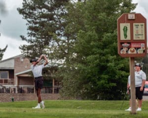 Young golfer swinging a club on the course, with fellow player and hole sign visible in the background.