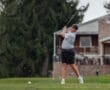 Young golfer swings on the course, with a house in the background and grass flying from the club striking the ball.