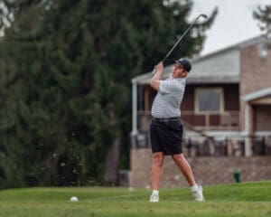 Young golfer swings on the course, with a house in the background and grass flying from the club striking the ball.