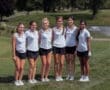 Six female golfers in matching uniforms pose together on a lush green golf course, smiling in a sunny outdoor setting.