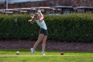 A golfer swings a club on the course, focusing on her technique with a lush green background.