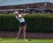 Young female golfer in action, swinging a club on the course with golf balls nearby and lush greenery in the background.
