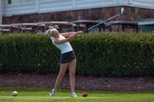 Young female golfer in action, swinging a club on the course with golf balls nearby and lush greenery in the background.