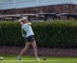 Young female golfer taking a swing on a green golf course, with golf carts and shrubbery in the background.