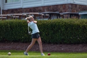 Young female golfer taking a swing on a green golf course, with golf carts and shrubbery in the background.