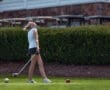 Young female golfer celebrating her swing on the course, with golf balls in front of her and carts in the background.