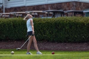 Young female golfer celebrating her swing on the course, with golf balls in front of her and carts in the background.