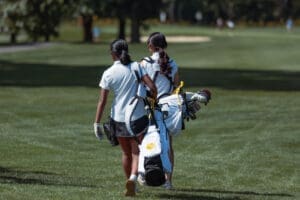 Two young girls walking on a golf course, carrying golf bags under sunny skies, showcasing youth sports enthusiasm.