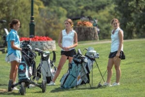 Three female golfers in casual attire gather on the course, discussing their game while surrounded by golf bags.