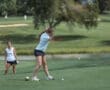 Two young women on a golf course; one tees off while another watches nearby, showcasing a beautiful golf setting.