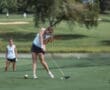 Young female golfer swinging a club on a green course, focused on her shot with another player in the background.