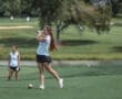 Young female golfer preparing to tee off, with a scenic course and another player in the background.