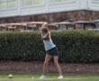 A young girl swings a golf club on a course, focused on her shot amidst green grass and a backdrop of bushes.