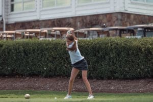 A young girl swings a golf club on a course, focused on her shot amidst green grass and a backdrop of bushes.