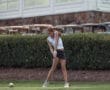 Young female golfer preparing to swing her club on a golf course, with lush greenery and golf carts in the background.