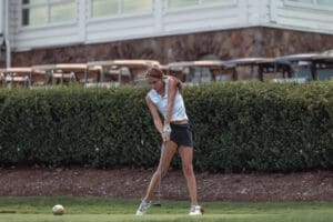 Young female golfer preparing to swing her club on a golf course, with lush greenery and golf carts in the background.