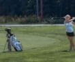 Young female golfer in action, swinging a club with her golf bag beside her on a sunny course.