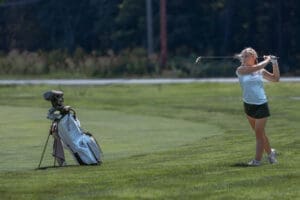 Young female golfer in action, swinging a club with her golf bag beside her on a sunny course.