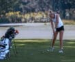 Young woman practicing her golf swing on the course, with golf clubs and bag in the background.