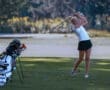 Young female golfer in mid-swing on the course, with golf clubs and bag in the background, showcasing athletic skill.