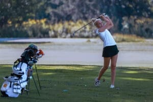 Young female golfer in mid-swing on the course, with golf clubs and bag in the background, showcasing athletic skill.