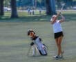 Young female golfer swings her club on a sunny day, with a golf bag and clubs resting nearby on the green.