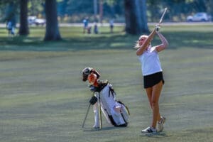 Young female golfer swings her club on a sunny day, with a golf bag and clubs resting nearby on the green.