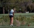 Young female golfer hitting a sand shot at a lush golf course, with a red ball nearby and flags in the background.