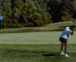 A golfer prepares to putt on a lush green course, aiming for the flag near the hole under clear blue skies.