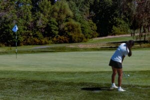 A golfer prepares to putt on a lush green course, aiming for the flag near the hole under clear blue skies.