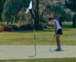 Young female golfer preparing to putt on a sunny day, with a flag in the background on a lush green golf course.