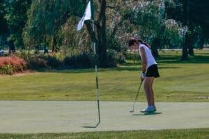 Young female golfer preparing to putt on a sunny day, with a flag in the background on a lush green golf course.