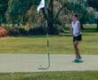 A young woman in a sleeveless shirt practices putting on a sunny golf course, with the flagstick in the background.