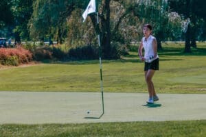 A young woman in a sleeveless shirt practices putting on a sunny golf course, with the flagstick in the background.