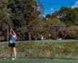 A young female golfer swings her club on a sunny day, aiming for the fairway with a red tee in the foreground.