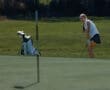Young golfer preparing to putt on a sunny day, with golf clubs and bag in the background on a well-maintained green.