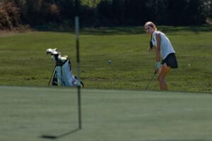 Young golfer preparing to putt on a sunny day, with golf clubs and bag in the background on a well-maintained green.