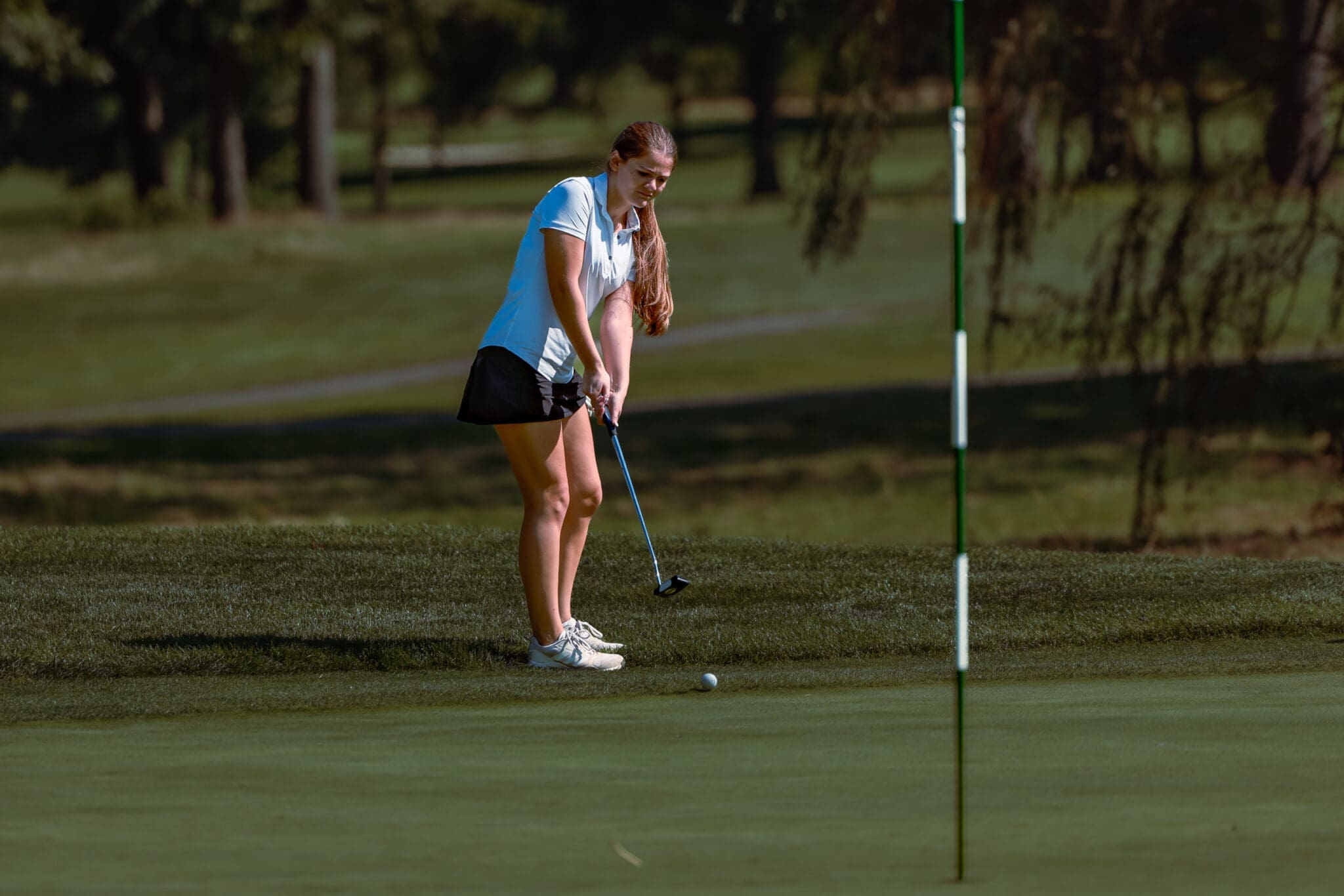 Female golfer in a white shirt and black skirt prepares to chip onto the green near the hole on a sunny day.