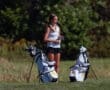 Young female golfer preparing for a game, standing beside two golf bags on a sunny course with greenery in the background.