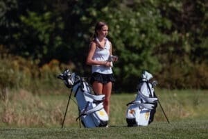 Young female golfer preparing for a game, standing beside two golf bags on a sunny course with greenery in the background.