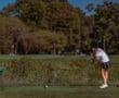 A golfer in a gray shirt and skirt swings at the ball on a lush golf course under a clear blue sky.