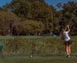 Female golfer tees off on a sunny day, surrounded by greenery and a scenic golf course.