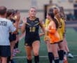 Girl in soccer uniform waves during a team lineup, coach encourages players in the background on the field.